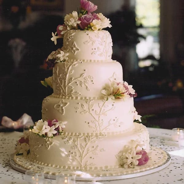 A four-tiered white wedding cake with intricate floral icing, silver beads, and fresh flowers.