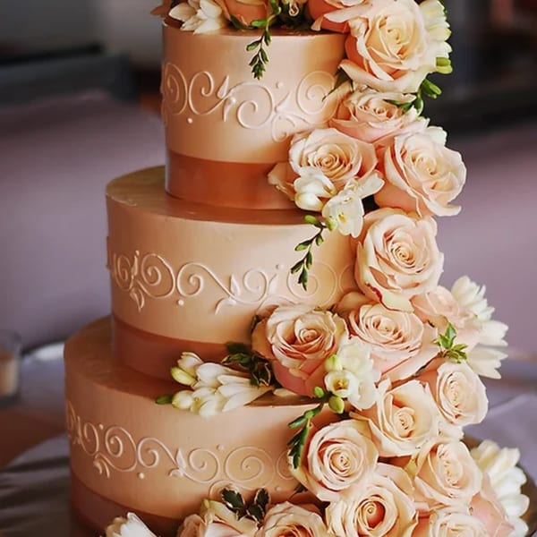 A three-tiered wedding cake with peach frosting, white scrollwork, and cascading blush roses.