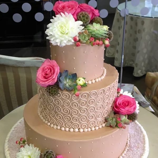 A three-tiered cake with pink frosting, decorated with pink roses, white flowers, and succulents.