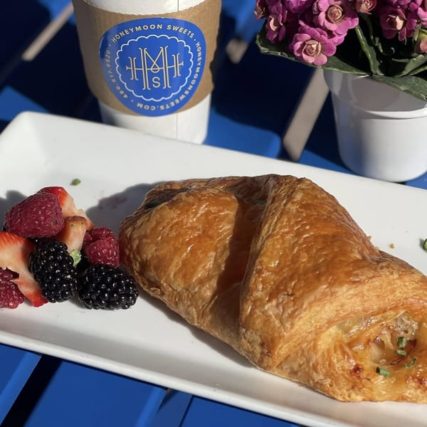 A golden pastry with berries on a white plate, a Honeymoon Sweets coffee, and purple flowers.