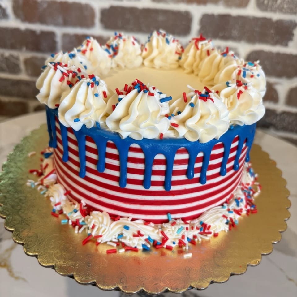 Red, white, and blue striped cake with blue icing drip and white frosting rosettes topped with sprinkles.