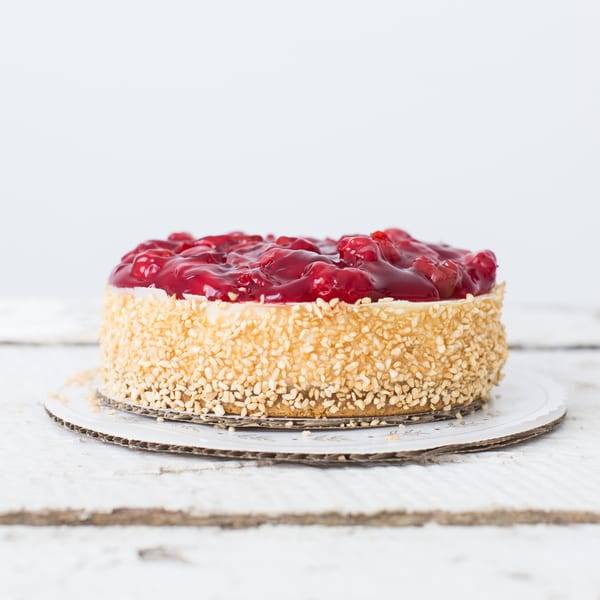 Honeymoon Sweets European Bakery's cheesecake with cherry topping and nut-covered sides on a white wooden surface.