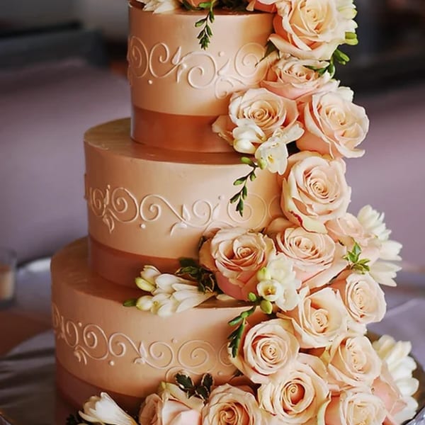 Three-tiered wedding cake with peach frosting, white scrollwork, and cascading peach roses and white flowers.