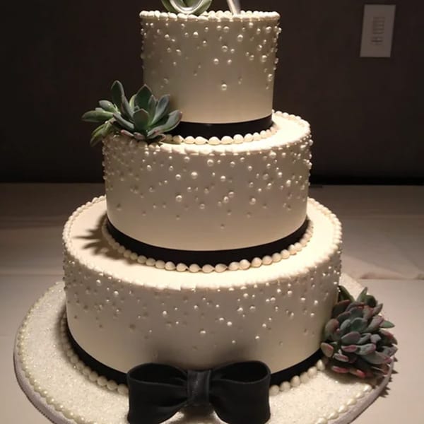 Three-tiered white wedding cake with black ribbons, white pearl accents, and succulent decorations.