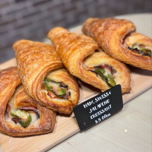 Four golden-brown ham, Swiss, and jalapeño croissants on a wooden board with a price tag.