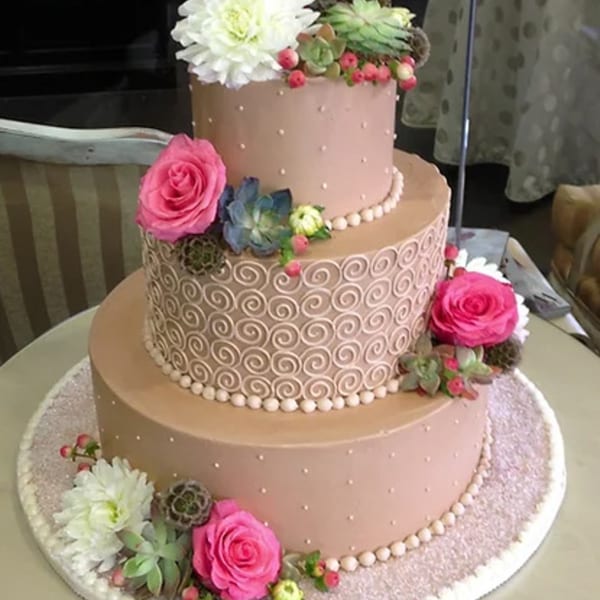 Three-tiered pink cake with white and pink flowers, green succulents, and white beaded and swirl frosting.