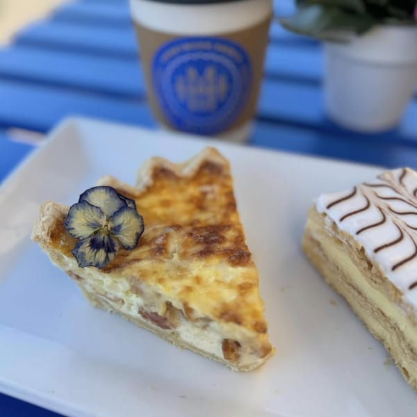 A slice of quiche with an edible flower and a pastry on a white plate with coffee.