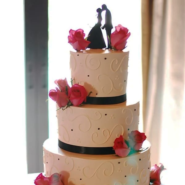 Three-tier wedding cake with black ribbons, pink roses, and a silhouetted bride and groom topper.