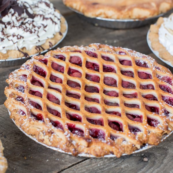 Close-up of a cherry lattice pie with powdered sugar, surrounded by other pies on a wooden table.