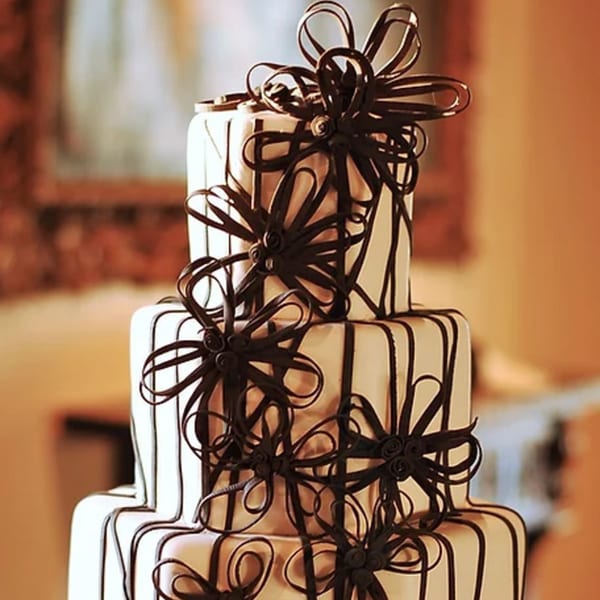 A multi-tiered wedding cake with white frosting and intricate dark chocolate ribbon decorations.