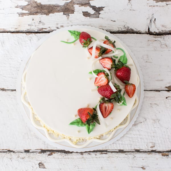 Round white cake with fresh strawberries, white chocolate shavings, and green icing leaves on a rustic white table.