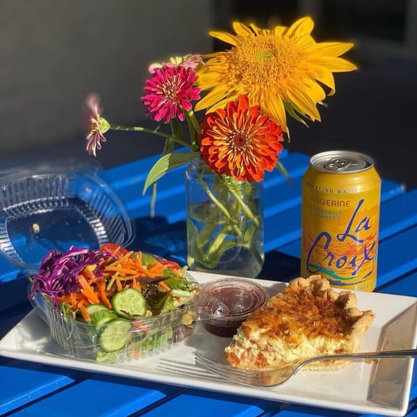 Quiche, side salad, La Croix, and a vase of flowers on a blue outdoor table.