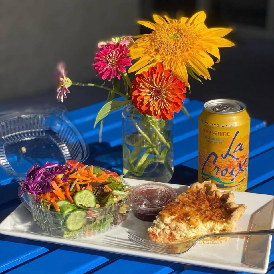 Quiche, side salad, La Croix, and a vase of flowers on a blue outdoor table.