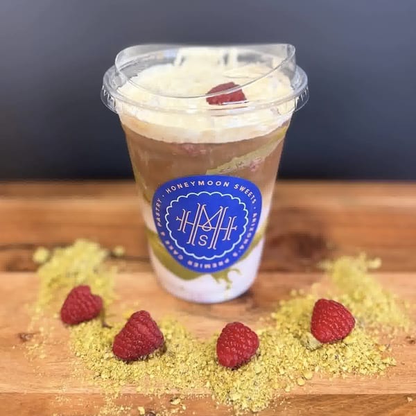 Honeymoon Sweets' layered dessert cup with raspberries and pistachio dust on a wooden surface.
